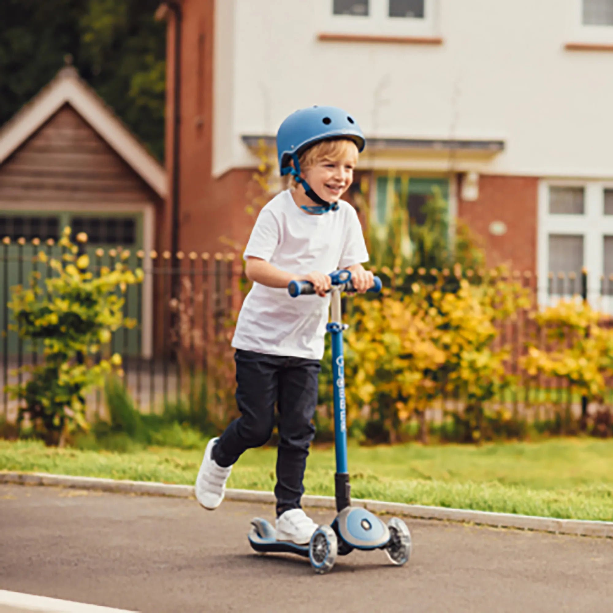 Smiling boy riding a neon pink elite deluxe foldable and adjustable 3 wheel scooter wearing a globber primo navy blue helmet.