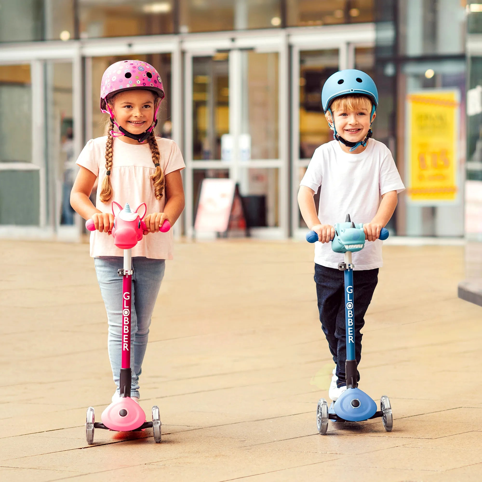 Boy riding a navy blue primo foldable 3 wheel scooter and girl riding a deep pink model. Both wearing helmets and smiling.